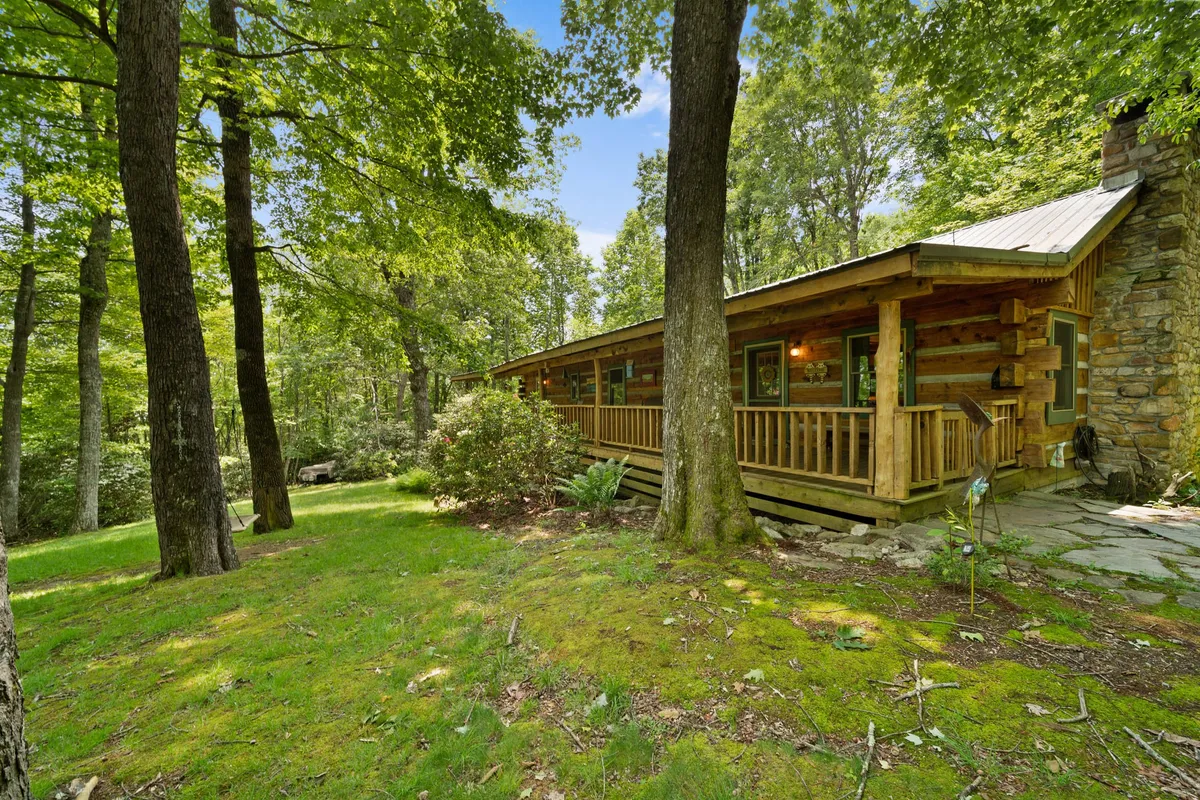 This image shows the log cabin structure with a prominent stone chimney, a spacious wooden porch, and lush greenery surrounding the home. A stone pathway leads up to the porch, evoking a sense of immersion in nature and providing a visual entryway for outdoor activities. The setting appears tranquil and inviting, perfect for relaxing after an adventure or appreciating the natural beauty of the area.