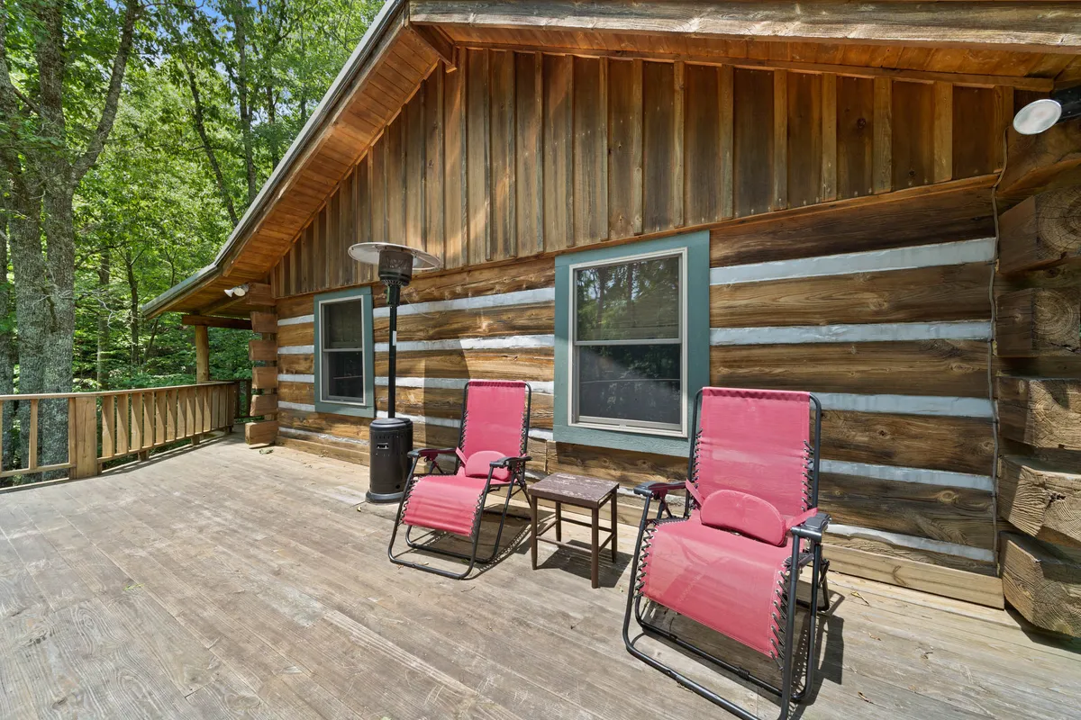 The image features the cabin’s exterior, with red reclining chairs arranged on a small deck alongside a side table and outdoor heater. The peaceful setting is surrounded by trees and greenery, suggesting a place to unwind, stargaze, or observe wildlife—activities tied to creekside relaxation and evening enjoyment after a day of adventure.