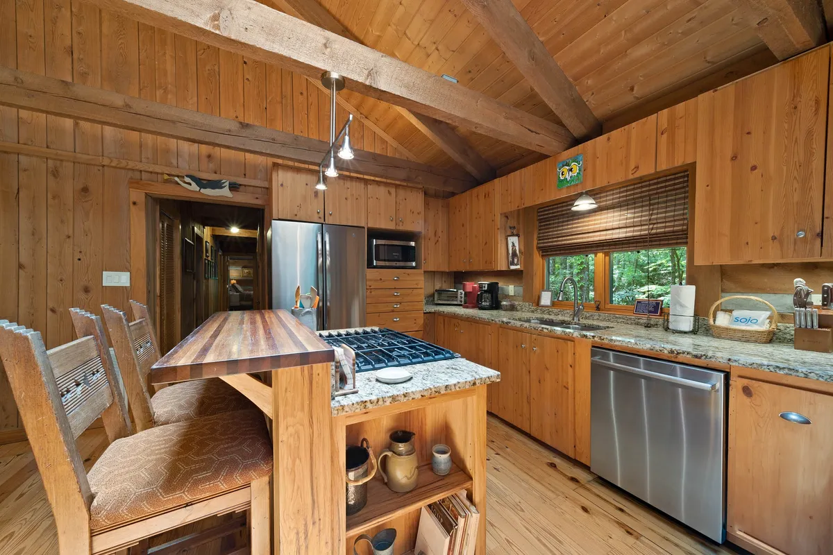 A rustic kitchen with granite countertops, warm wooden cabinetry, bar seating, and modern appliances. A window offers woodland views, and the kitchen is open, making it easy for chefs to socialize with guests in adjacent areas.