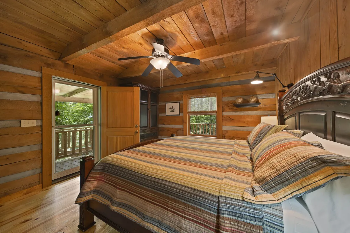 This image features a cozy bedroom with a colorful striped blanket on the bed, wooden ceiling and walls, and an open door leading to a balcony. The room is bathed in natural light, creating a warm and inviting atmosphere that's perfect for winding down after a day of adventure.