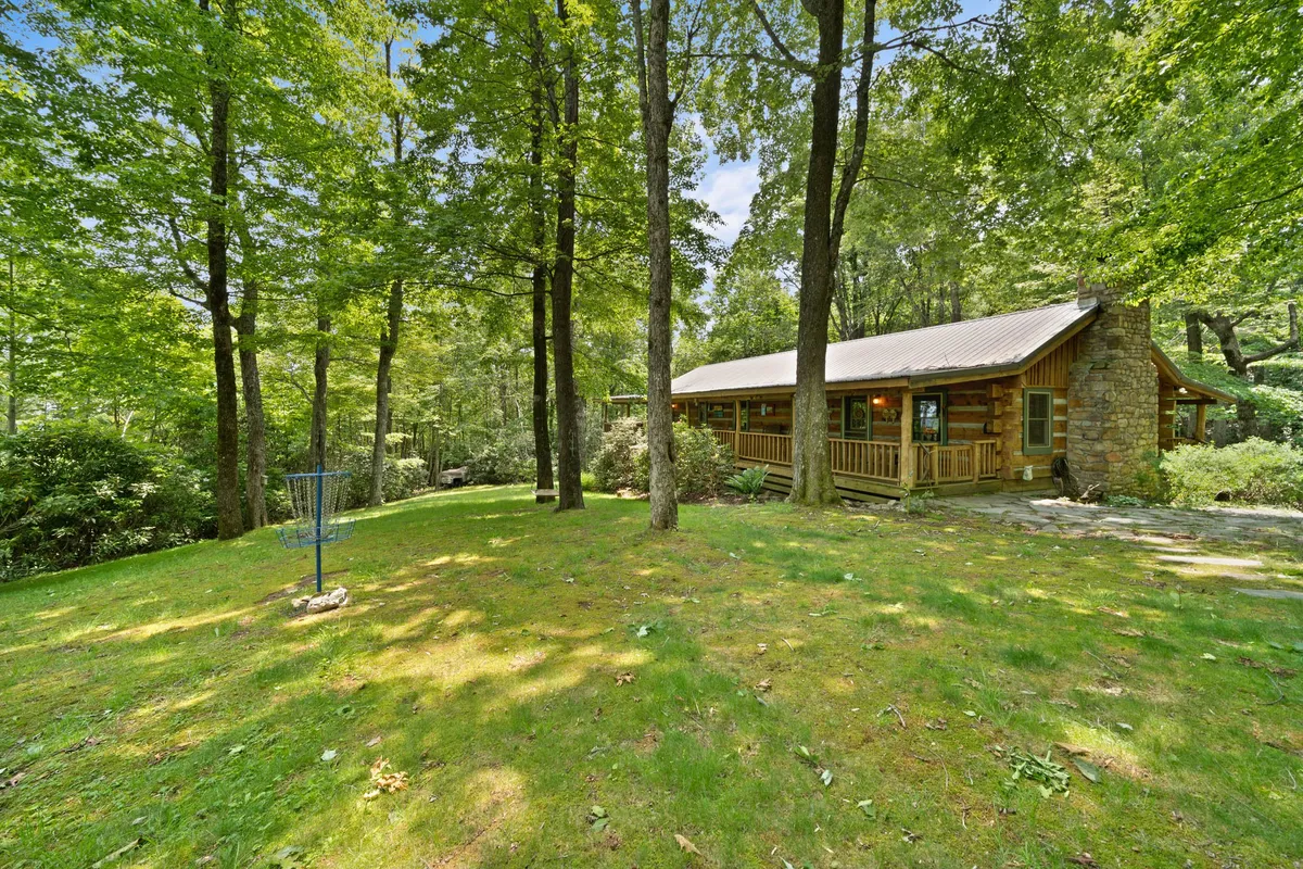 A picturesque log cabin is nestled among tall trees, with a welcoming front porch and a spacious lawn area. The porch features comfortable seating, perfect for relaxing after a day of adventure. The natural surroundings evoke a tranquil atmosphere that invites families to step outside and explore.