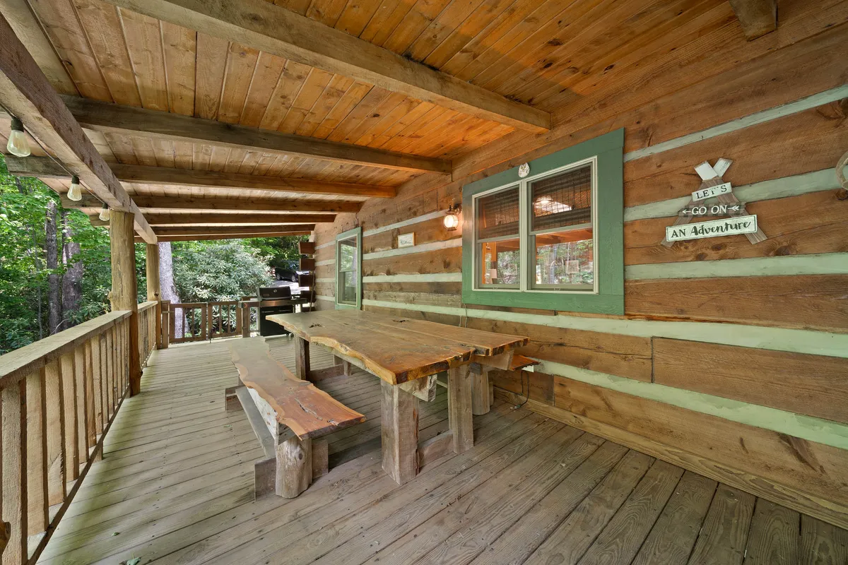 This image features a wooden table set on a porch against the log cabin walls, with green window trim visible. The table setting is ideal for outdoor dining and gathering, and the scene is surrounded by lush greenery, creating a sense of being immersed in nature.