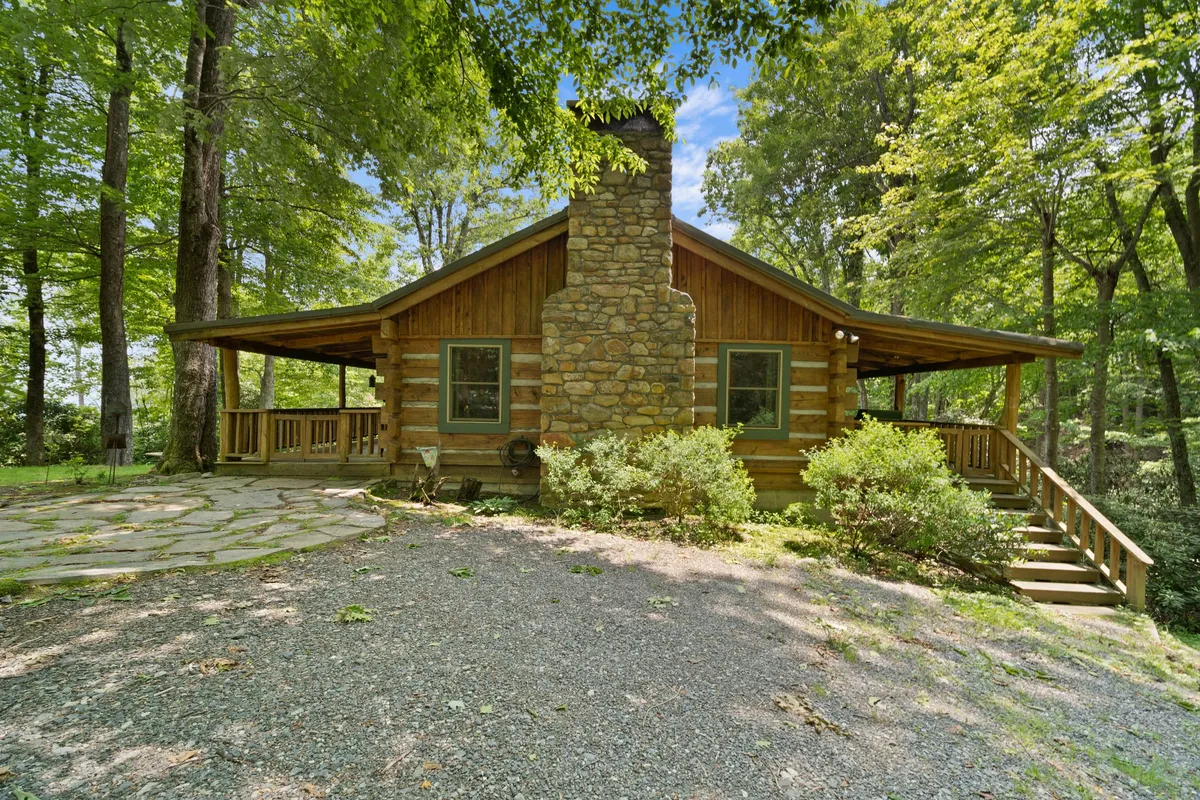 The exterior hero image showcases a classic log cabin nestled among towering trees, featuring a sturdy stone chimney, inviting wooden deck, and a tranquil creek just beyond the porch. The cozy porch suggests a perfect spot for early morning coffee, while the surrounding forest promises privacy and connection with nature.