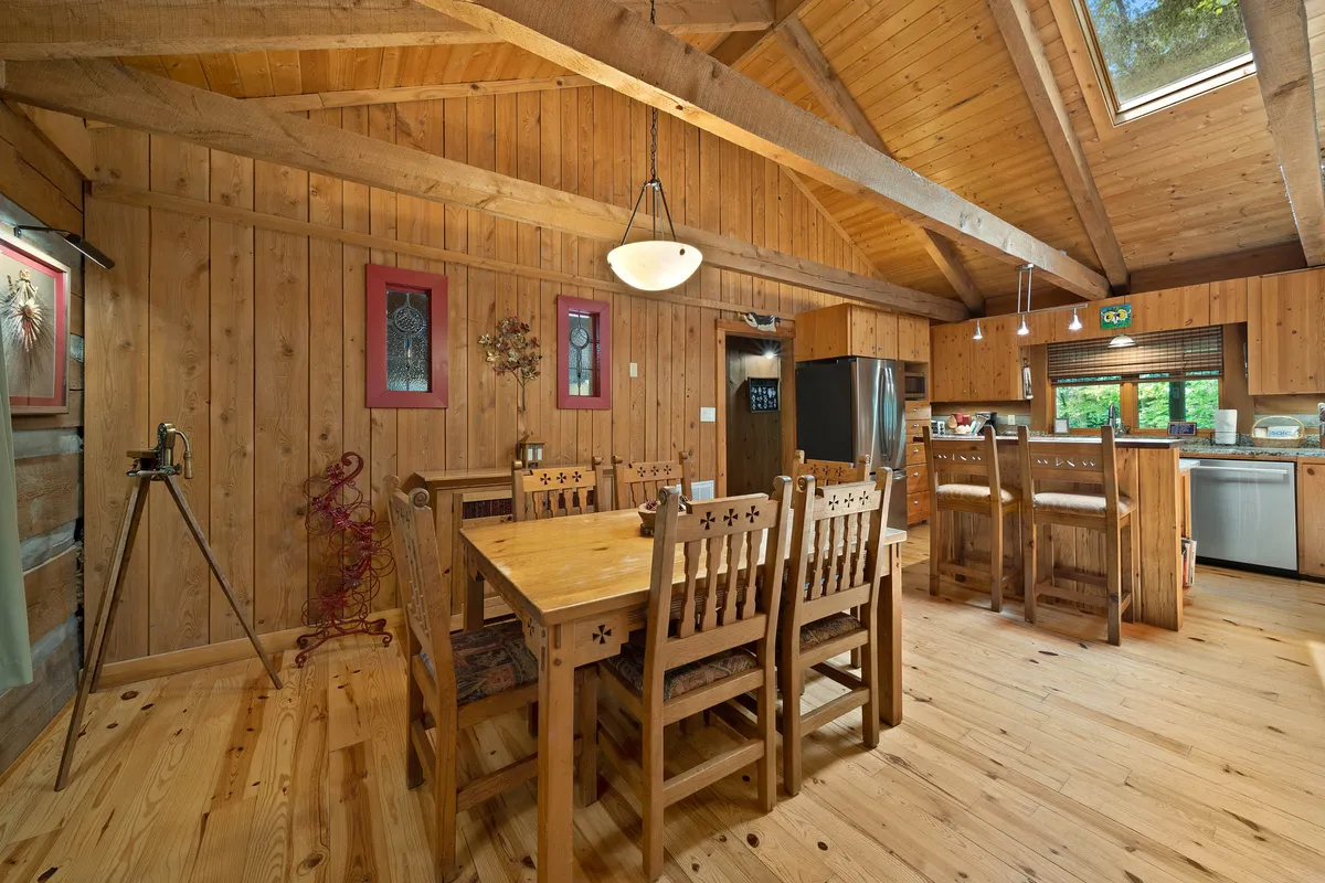 An inviting dining area featuring a long wooden table beneath high ceilings with exposed beams. The kitchen is visible in the background, and decorative wall art adds warmth and personality to the space, encouraging family meals and gatherings.