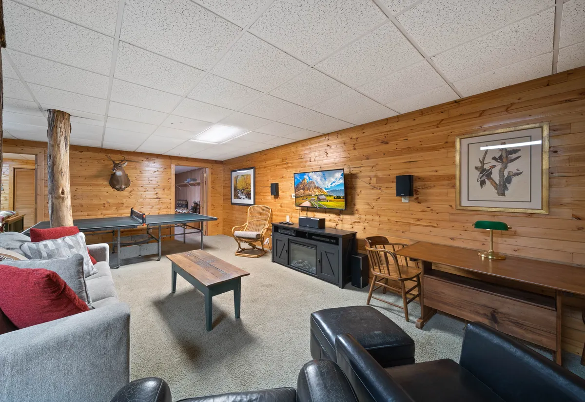 This image captures a vibrant game room featuring a ping pong table as the focal point. A cozy sofa with red cushions sits nearby, facing a flat screen television mounted on the wall. Wooden wall art and a desk with a lamp contribute to the warm, inviting ambiance. The scene suggests multiple activities taking place: a friendly ping pong match, relaxing on the sofa, or watching a movie.