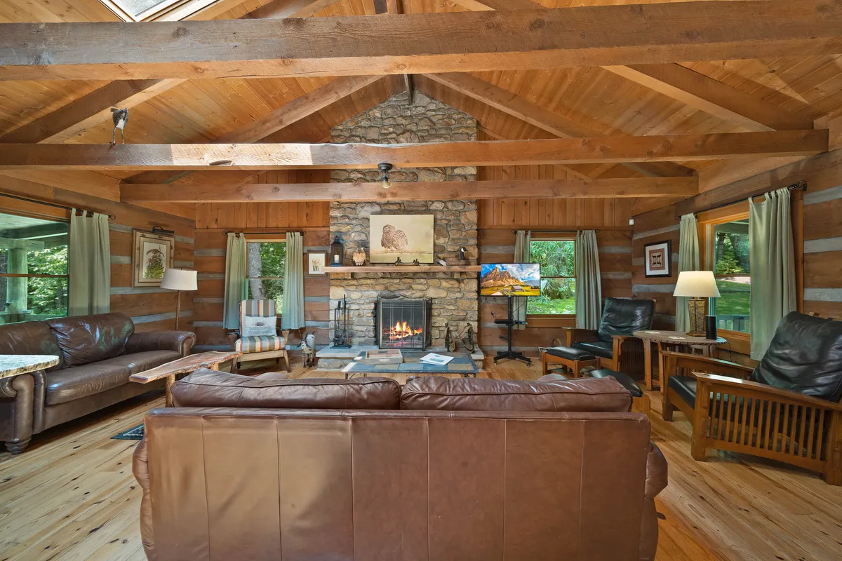 A spacious living room featuring a dramatic two-story stone fireplace, exposed wooden beams, and leather sofas arranged for group conversation. Large windows allow natural light to fill the space, creating a warm and welcoming atmosphere ideal for gathering and relaxing.