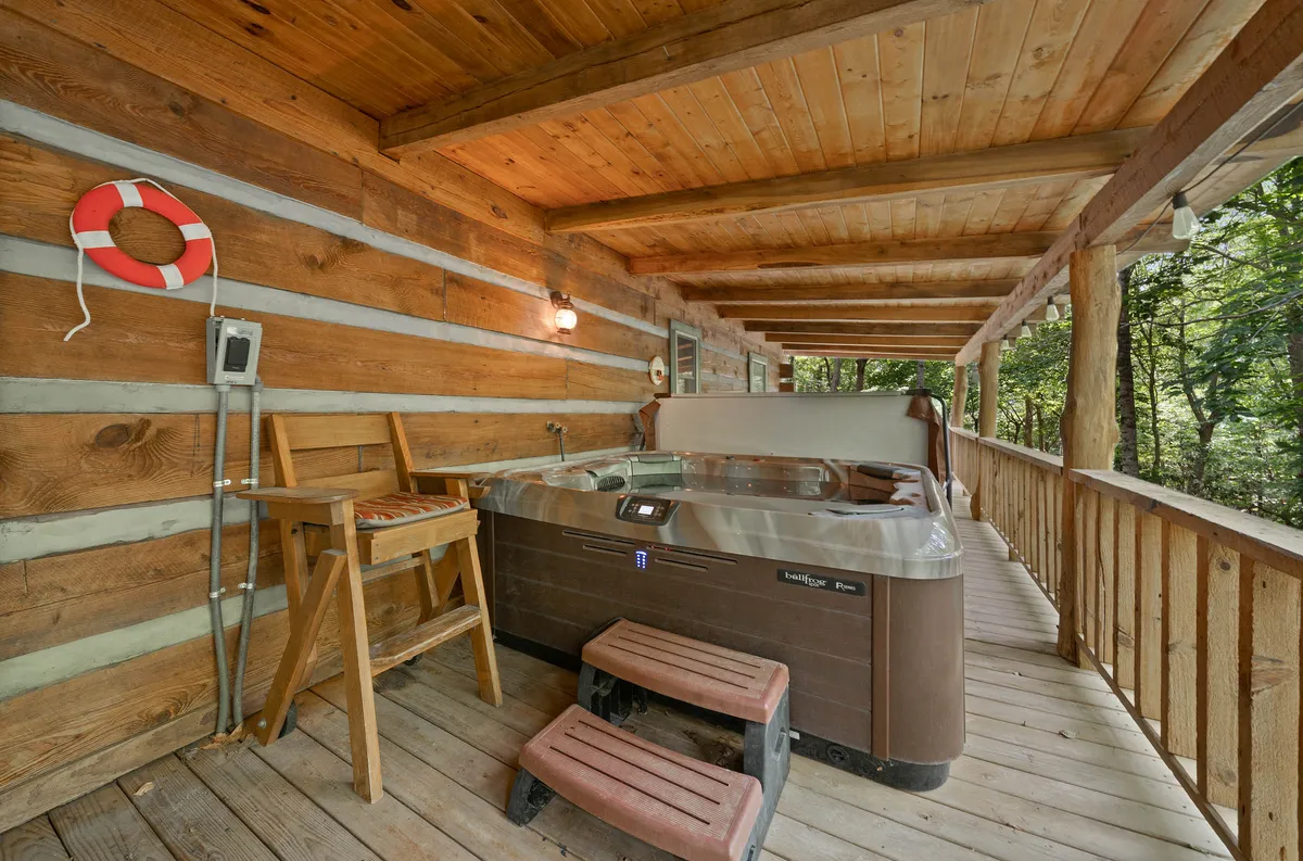 A spacious wooden deck with a large hot tub as the focal point, surrounded by natural greenery. A wooden chair is nearby, and the setup overlooks trees, offering privacy and tranquility. The hot tub is perfectly positioned for both soaking and enjoying the natural surroundings, with enough space for several people.