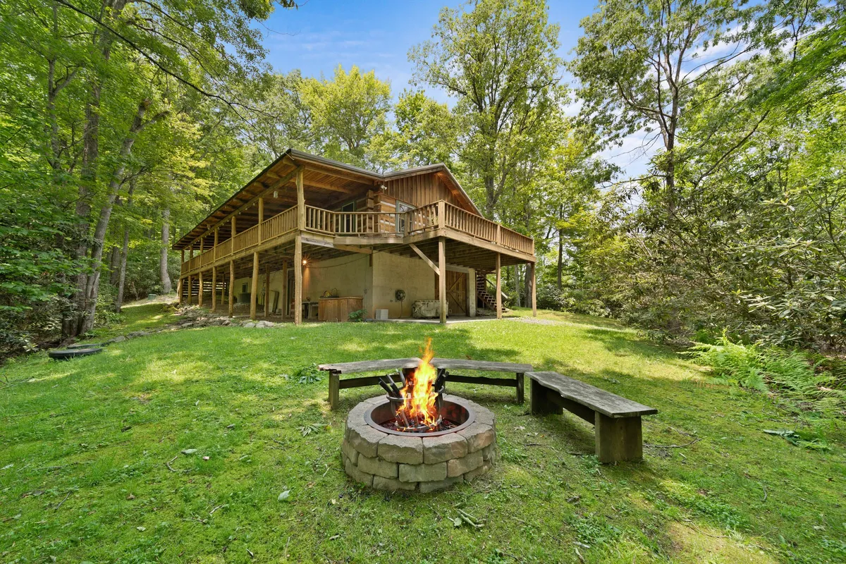 An inviting creekside area featuring a fire pit encircled by wooden benches and a nearby deck. The fire pit is the central gathering spot, framed by surrounding trees and the sound of nearby water, ideal for evening campfire gatherings, marshmallow roasting, and stargazing.
