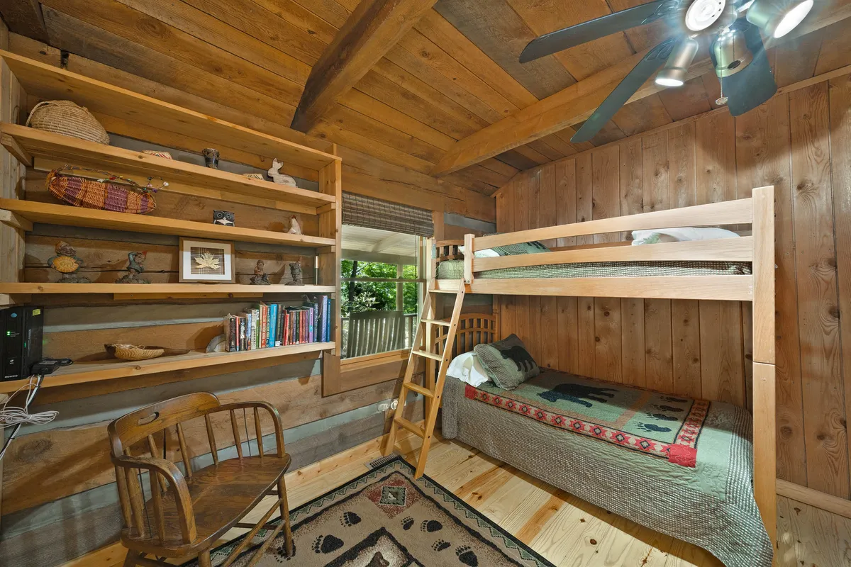 The image shows a playful and comfortable bunk room featuring wooden bunk beds, a bookshelf filled with books, a wooden chair, and a window with a view to the outdoors. The room feels inviting and designed with children or young guests in mind.